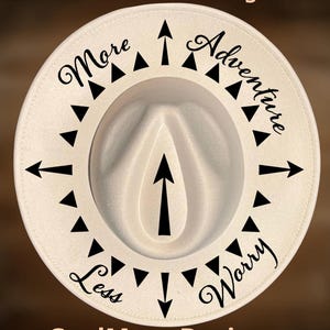 May include: Cream-colored fedora hat with a wide brim. The hat features black text that reads "More Adventure Less Worry" and "For Hand Burning". Black arrows and triangles decorate the brim. The text "CoolMoonDesigns" is at the bottom.