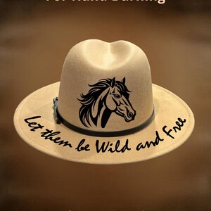May include: A tan fedora hat featuring a black horse head graphic and the text "Let them be Wild and Free" on the brim. The hat includes a thin black band around the crown. The text "For Hand Burning" and "CoolMoonDesigns" appear in the image.