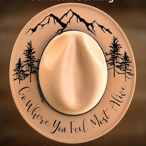 May include: A tan felt hat featuring black graphic designs including a mountain range at the top, pine trees on the sides, and the text "Go Where You Feel Most Alive" in a cursive script. The text "For Hand Burning" and "CoolMoonDesigns" appear.
