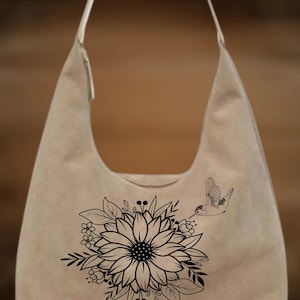 Puede incluir: Bolso de hombro beige con un diseño floral y colibrí negro. El bolso presenta un gran girasol con hojas y pequeñas flores, y un colibrí. Tiene una correa larga.