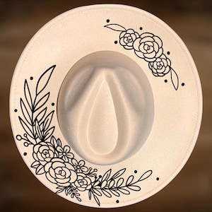Op de afbeelding: Een crèmekleurige fedora hoed met een brede rand. De rand is versierd met een zwart bloemenontwerp. De hoed heeft een druppelvormige kroon en een grosgrain lint.