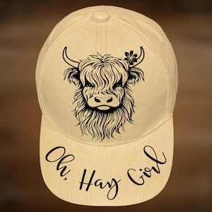May include: A beige baseball cap featuring a black graphic illustration of a Highland cow with a small flower on its head. The text "Oh, Hay Girl" is printed in black script on the brim. The text "For Hand Burning" and "CoolMoonDesigns" appear.