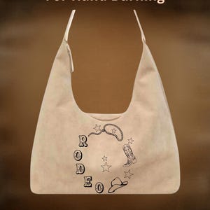 May include: Beige hobo bag with a black rodeo-themed design. The design includes the word "RODEO", a lasso, stars, a cowboy boot, and a cowboy hat. The text "For Hand Burning" and "CoolMoonDesigns" are also visible.