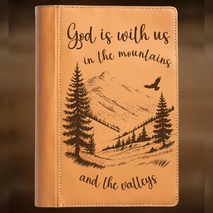 May include: Tan leather journal with the text "God is with us in the mountains and the valleys." The cover features a hand-burned mountain and tree design, with an eagle soaring above. The words "For Hand Burning" and "CoolMoonDesigns" are also visible.