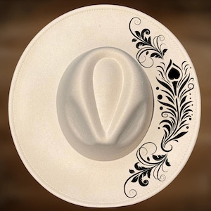 May include: Cream-colored fedora hat with a decorative black floral design on the side. The hat has a wide brim and a structured crown. The hat is a stylish accessory.