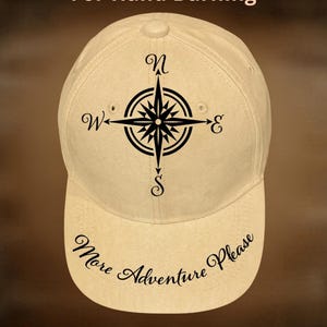 May include: A cream-coloured baseball cap featuring a black compass rose emblem. The brim displays the text "More Adventure Please". The cap also has the text "For Hand Burning" and "CoolMoonDesigns".