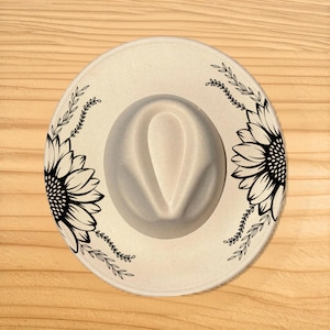 Puede incluir: Sombrero fedora color crema con corona gris. El sombrero presenta un diseño de girasol negro a cada lado del ala, con detalles de hojas adicionales. Un accesorio elegante.