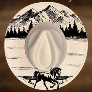 Op de afbeelding: Een crèmekleurige cowboyhoed met een zwart berg- en bosontwerp rond de rand. Een zwarte paardenafbeelding staat op de voorkant. De tekst "For Hand Burning" en "CoolMoonDesigns" zijn ook zichtbaar.