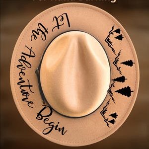 May include: Tan fedora-style hat with a black band. The hat features the text "Let the Adventure Begin" and a mountain and tree design. The words "For Hand Burning" and "CoolMoonDesigns" are also visible.