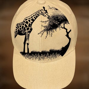 May include: A beige baseball cap featuring a black and white graphic of a giraffe reaching up to eat leaves from a tree. The giraffe has a distinctive spotted pattern. The cap has a curved brim and a button on top.