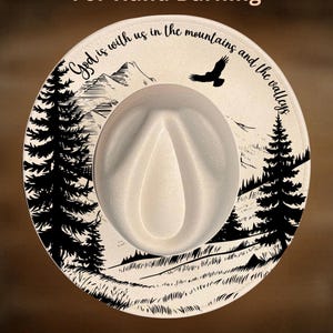 May include: Cream-colored fedora hat with a black and white landscape design. The hat features a mountain scene, trees, and a flying bird, with the text "God is with us in the mountains and the valleys". The text "For Hand Burning" and "CoolMoonDesigns" are also visible.