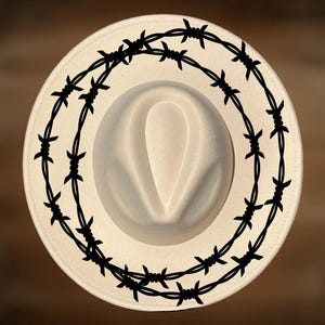 Puede incluir: Un sombrero fedora color crema con un diseño de alambre de púas negro que rodea el ala. El sombrero tiene una corona clásica en forma de lágrima y un ala ancha. Un accesorio elegante.