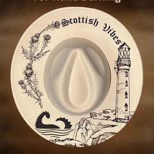 May include: Cream-colored fedora hat with a Scottish theme. The hat features black illustrations of a thistle, lighthouse, and the Loch Ness Monster. Text on the hat includes "Scottish Vibes", "For Hand Burning", and "CoolMoonDesigns".