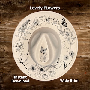 May include: Cream-colored fedora hat with a wide brim, adorned with black floral and butterfly illustrations. The hat features the text "Lovely Flowers" and "Instant Download" and "Wide Brim". The hat is set against a wooden background.