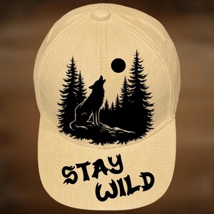 Può includere: Un cappellino da baseball color marrone chiaro con una grafica nera di un lupo che ulula alla luna tra alberi di pino. Il testo "STAY WILD" è stampato sotto la grafica. Il testo "For Hand Burning" appare in alto e "CoolMoonDesigns" in basso.