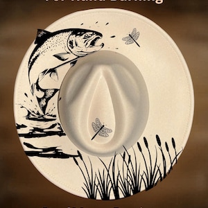 Pode incluir: Chapéu fedora creme com um gráfico preto de um peixe saltando, libélulas e juncos. O texto "For Hand Burning" e "CoolMoonDesigns" está impresso no chapéu.