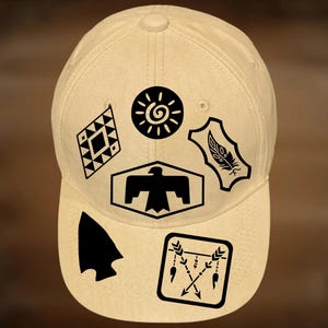 Puede incluir: Una gorra de béisbol beige con estampados gráficos negros de un sol, un patrón de diamante, una pluma, la silueta de un pájaro, una punta de flecha y flechas cruzadas. El texto For Hand Burning y CoolMoonDesigns está impreso en la gorra.