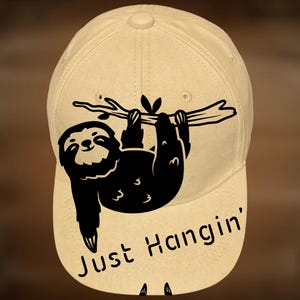 May include: A beige baseball cap with a black sloth graphic hanging from a branch. The text "Just Hangin'" is below the sloth, and "For Hand Burning" and "CoolMoonDesigns" are above and below the sloth, respectively.