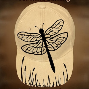 May include: A beige baseball cap featuring a black dragonfly design. The cap has the text "For Hand Burning" and "CoolMoonDesigns" printed on it. The dragonfly is centered on the front of the cap.