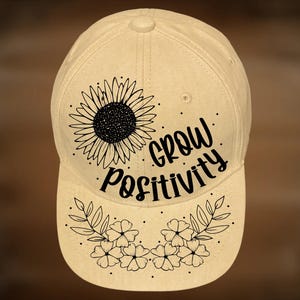 May include: A beige baseball cap with a sunflower and floral design. The cap features the words "Grow Positivity" in a handwritten font. The text "For Hand Burning" and "CoolMoonDesigns" are also visible.