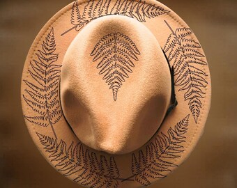 Puede incluir: Un sombrero fedora de fieltro color canela con diseños de hojas de helecho pirograbados en negro en la copa y el ala. El texto "For Hand Burning" aparece en la parte superior y "CoolMoonDesigns" en la inferior. Tiene una copa estructurada.