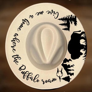 May include: Cream-colored fedora hat with a wide brim. The hat features black graphic designs of trees, a bison, and text that reads "Give me a home where the Buffalo roam." The text "For Hand Burning" and "CoolMoonDesigns" are also visible.