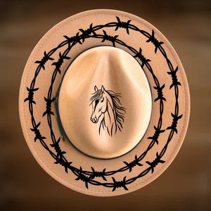 Puede incluir: Sombrero de vaquero color beige con un diseño de alambre de púas negro alrededor del borde. El centro del sombrero presenta una ilustración negra de la cabeza de un caballo. El sombrero tiene una forma de fedora clásica.
