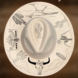 May include: A cream-colored fedora hat with black hand-drawn desert-themed illustrations. The hat features a central cactus design and a brim adorned with cacti, a roadrunner, and a skull with floral accents. The text "For Hand Burning" and "CoolMoonDesigns" are also visible.
