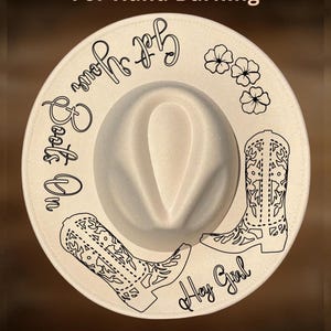 Può includere: Un cappello fedora color crema con tesa larga. Il cappello presenta disegni a tratto nero di stivali da cowboy, fiori e le frasi "Get Your Boots On" e "Hey Girl". È visibile anche il testo "For Hand Burning" e "CoolMoonDesigns".