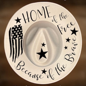May include: Cream-colored fedora hat with the text "HOME of the Free Because of the Brave." The hat features a black and white American flag design and black star accents. The text "For Hand Burning" and "CoolMoonDesigns" are also visible.