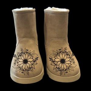May include: A pair of tan suede boots with a black floral design on the back of each boot.