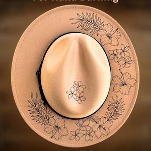 Op de afbeelding: Beige fedora hoed met zwarte bloempatronen en de tekst "For Hand Burning" en "CoolMoonDesigns". De hoed heeft een zwarte band en een afgeronde kroon. De bloemmotieven omvatten hibiscus en andere tropische bloemen.