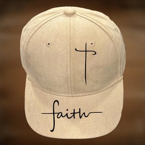 May include: A beige baseball cap with a curved brim. The cap features a black cross design on the front and the word "faith" in a script font below the cross. The cap is made of a soft, textured material.
