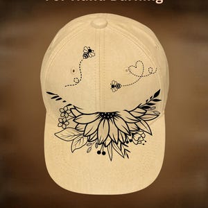 May include: Cream-colored baseball cap with a black floral and bee design. The cap features a sunflower and leaf design on the front, with two bees and a heart. The text "For Hand Burning" and "CoolMoonDesigns" are also visible.