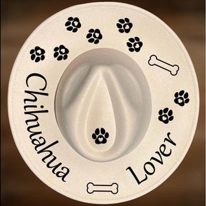 Puede incluir: Un sombrero fedora color crema con una corona blanca, con huellas de patas negras y gráficos de huesos. El sombrero está inscrito con "Chihuahua Lover", "For Hand Burning" y "CoolMoonDesigns".