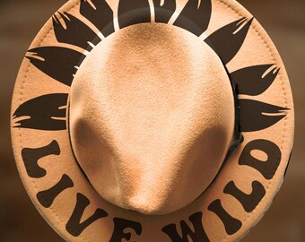 May include: A tan felt fedora hat featuring a black sunflower petal design around the crown and the text "LIVE WILD" printed in bold black lettering along the brim. The hat has a structured crown and a wide, flat brim with visible stitching.