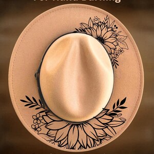 May include: A tan fedora-style hat with a black floral design and the text "For Hand Burning" and "CoolMoonDesigns". The hat has a black band and a rounded crown.