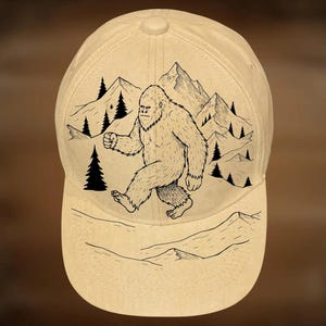 May include: A beige baseball cap featuring a black and white illustration of a Bigfoot walking in front of mountains and trees. The text "For Hand Burning" and "CoolMoonDesigns" are printed on the cap.