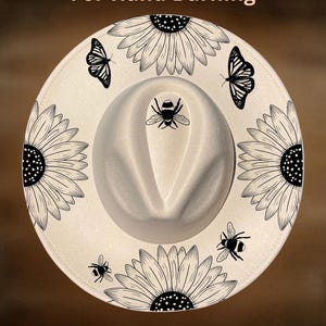May include: A cream-colored fedora hat featuring black line art illustrations of sunflowers, butterflies, and bees. The text "For Hand Burning" appears at the top and "CoolMoonDesigns" at the bottom. The hat has a pinched crown and a wide brim.