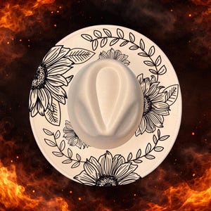 May include: Cream-colored fedora hat with a wide brim. The brim is decorated with black line art of sunflowers and leaves. The hat has a rounded crown and a neutral color scheme, suitable for various outfits.