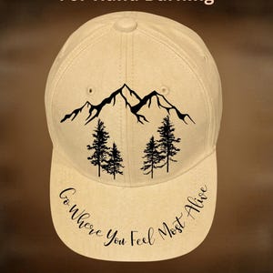 May include: Tan baseball cap with a black mountain and tree design. The text "Go Where You Feel Most Alive" is on the brim. The text "For Hand Burning" and "CoolMoonDesigns" are also visible.