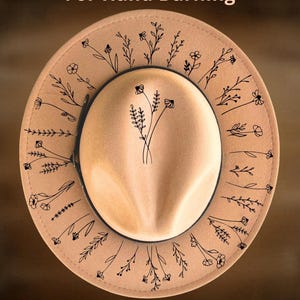 May include: A tan fedora hat featuring black line art of wildflowers on the brim and crown. The text "For Hand Burning" appears at the top and "CoolMoonDesigns" at the bottom. The hat has a wide brim and a pinched crown design.