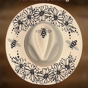 Può includere: Un cappello fedora bianco con un motivo floreale nero decorativo sul bordo e tre illustrazioni di api nere. Il testo "For Hand Burning" e "CoolMoonDesigns" è stampato sul cappello.