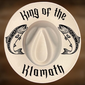 May include: A cream-colored fedora hat with the text "King of the Klamath" in black gothic font. Two black and white fish illustrations flank the hat's center. The text "For Hand Burning" and "CoolMoonDesigns" are also visible.