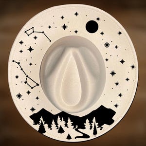 Può includere: Cappello fedora color crema con un design di montagna e foresta nera sul bordo. Il bordo presenta anche un motivo di costellazioni e stelle, con una luna nera. Il cappello ha una corona a goccia.