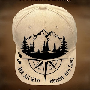 May include: A beige baseball cap with a black mountain and forest graphic. The cap features a compass design with the text "Not All Who Wander Are Lost." The text "For Hand Burning" and "CoolMoonDesigns" are also visible.