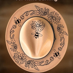 Può includere: Un cappello in feltro color marrone chiaro con un disegno a linee nere di api e margherite sulla corona e sulla tesa. Il testo "For Hand Burning" appare in alto e "CoolMoonDesigns" in basso. Il cappello ha una fascia scura alla base.