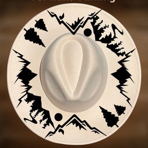 Può includere: Un cappello fedora color crema con una stampa grafica nera di catene montuose, pini e forme circolari sulla tesa. Il testo "For Hand Burning" appare in alto e "CoolMoonDesigns" è stampato nella parte inferiore.