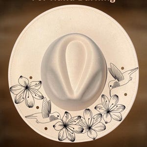 Puede incluir: Sombrero fedora color crema con corona redondeada y ala ancha. El ala está decorada con dibujos lineales negros de flores y colibríes. El texto "For Hand Burning" y "CoolMoonDesigns" está impreso en el sombrero.