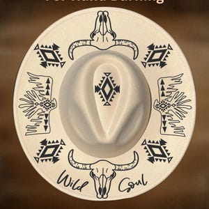 May include: Cream-coloured fedora hat with a wide brim, featuring black tribal and skull designs. The hat has the text "For Hand Burning", "Wild Soul", and "CoolMoonDesigns".